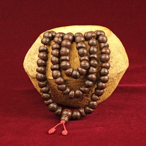 May include: A brown wooden mala bead necklace with a red string tassel. The necklace is wrapped around a light brown ceramic bowl.