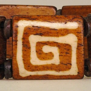 Puede incluir: Un brazalete de madera con cuadrados marrones y blancos. Cada cuadrado tiene un dise&ntilde;o de espiral blanco.