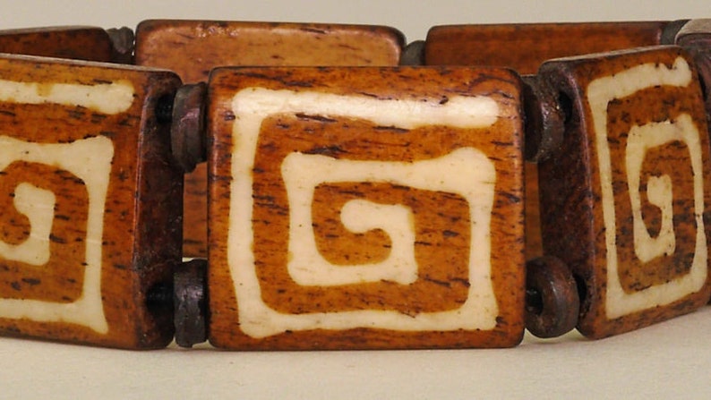 Puede incluir: Un brazalete de madera con cuadrados marrones y blancos. Cada cuadrado tiene un dise&ntilde;o de espiral blanco.