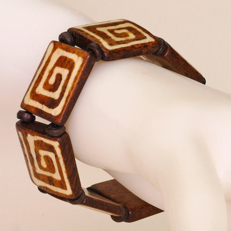 Puede incluir: Un brazalete de madera marr&oacute;n con dise&ntilde;os geom&eacute;tricos blancos. El brazalete est&aacute; hecho de varias piezas cuadradas que est&aacute;n conectadas entre s&iacute;.