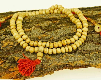 May include: A white wooden mala bead necklace with red tassels. The necklace is made of round beads and is strung on a cord. The necklace is lying on a piece of tree bark.