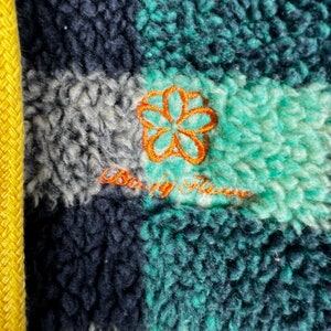 Op de afbeelding: Close-up van een groene, zwarte en witte geruite fleecejas met een oranje geborduurde bloem en de tekst "Baby, it's cold outside".