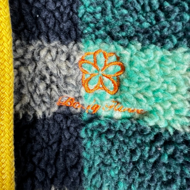 Op de afbeelding: Close-up van een groene, zwarte en witte geruite fleecejas met een oranje geborduurde bloem en de tekst "Baby, it's cold outside".