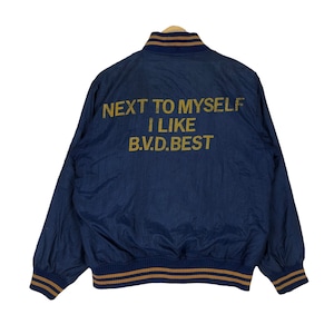 Könnte beinhalten: Eine marineblaue Bomberjacke mit braunem und goldenem geripptem Kragen und Bündchen. Auf der Rückseite der Jacke steht in goldenen Buchstaben "NEXT TO MYSELF I LIKE B.V.D.BEST".
