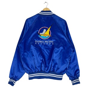 Peut inclure: Une veste bomber en satin bleu avec un col et des poignets rayés blanc et bleu. Le dos de la veste présente un logo brodé pour Global Pacific Records, qui comprend un marlin jaune sautant hors de l'eau.