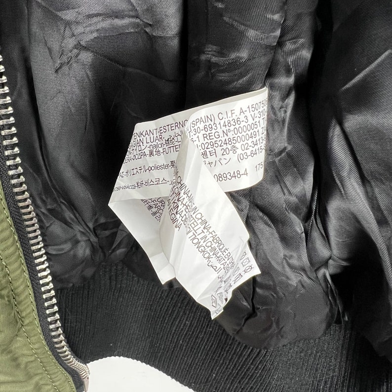 Puede incluir: Una chaqueta bomber verde con forro negro y cierre de cremallera. La etiqueta en el interior de la chaqueta tiene el siguiente texto: "ENKANT ESTERNO SPAIN) C.I.F. A-1507501:30-69314836-3 V-31 GAN LUAR الخارجي 9-1 REG.N&ordm;:0000051 T 1:029524850004912 센터 20층 02-3413 ジャパン (03-6415 089348-4 175"