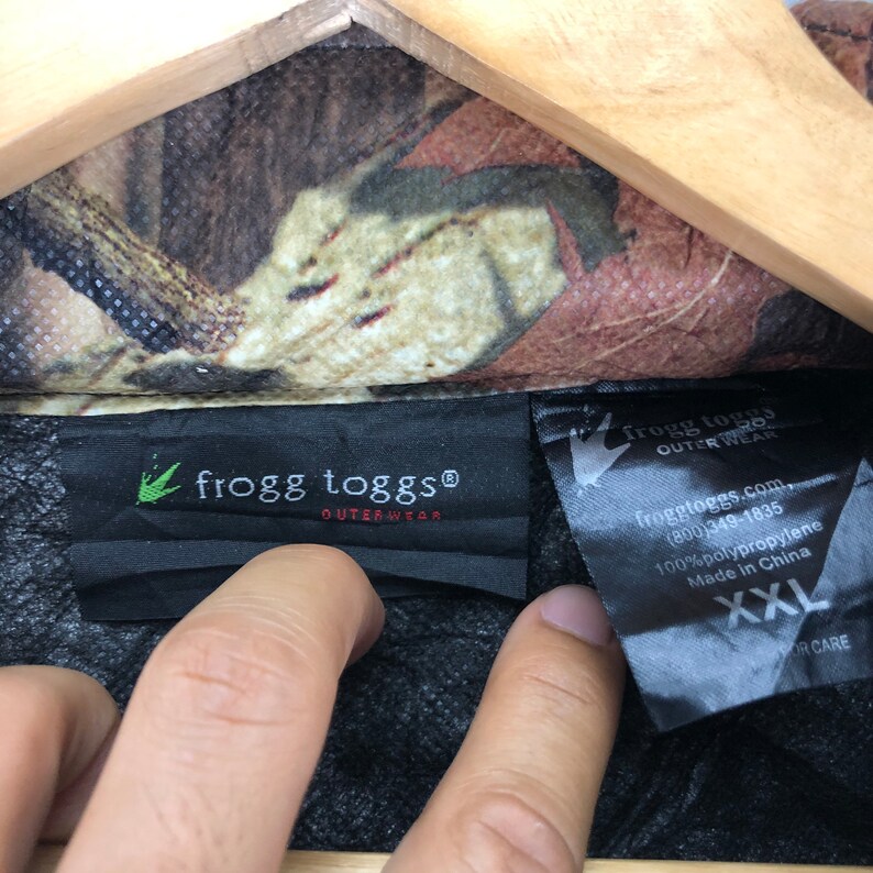 May include: A black and green camouflage jacket with a black label that reads "frogg toggs&reg; OUTERWEAR" and a smaller label that reads "frogg toggs OUTER WEAR froggtoggs.com (800)349-1835 100% polypropylene Made in China XXL OR CARE".