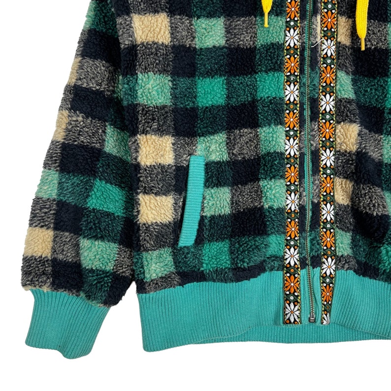 Op de afbeelding: Een groene, zwarte en cr&egrave;me geruite fleecejas met een turquoise rits en een rits met een bloemenpatroon. De jas heeft een turquoise geribbelde zoom en manchetten.