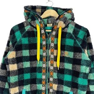 Op de afbeelding: Een groene, zwarte en cr&egrave;me geruite fleece hoodie met een gele trekkoord en een rits met een bloemenpatroon. De hoodie heeft een capuchon en een voorzak.