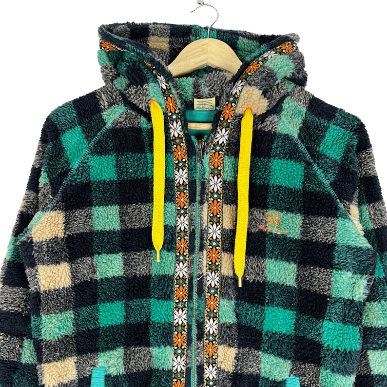 Op de afbeelding: Een groene, zwarte en cr&egrave;me geruite fleece hoodie met een gele trekkoord en een rits met een bloemenpatroon. De hoodie heeft een capuchon en een voorzak.