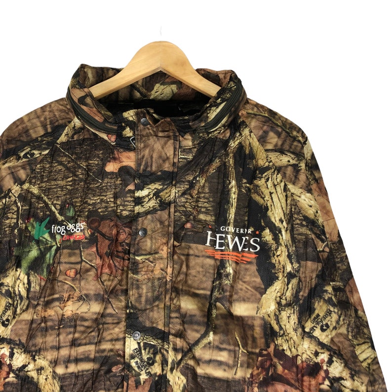May include: A camouflage jacket with a black zipper and a patch that says "GOVERNOR HEWES" in orange and white text. The jacket also has a green patch with a bird and the text "FROG OGGS" in white.