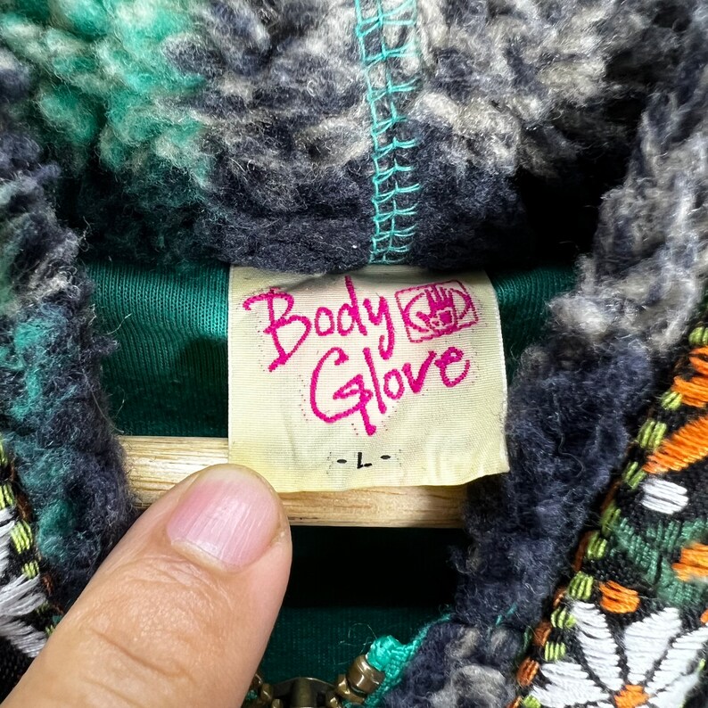 Op de afbeelding: Een groen en zwart fleecejack met een Body Glove-label. Het label is wit met roze tekst en een logo van een vuist.