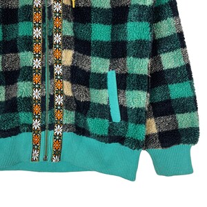Op de afbeelding: Een groen en zwart geruit fleecejack met een ritssluiting en een ritstrekker met bloemenprint. Het jack heeft een teal gekleurde zak aan de linkerkant en een teal gekleurde geribbelde zoom.