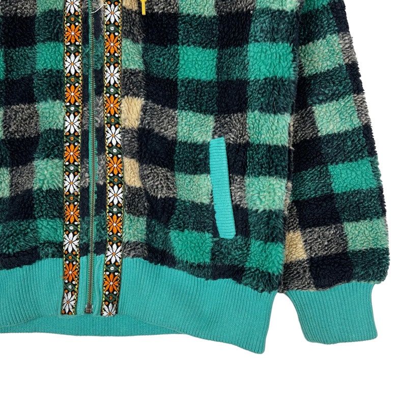Op de afbeelding: Een groen en zwart geruit fleecejack met een ritssluiting en een ritstrekker met bloemenprint. Het jack heeft een teal gekleurde zak aan de linkerkant en een teal gekleurde geribbelde zoom.
