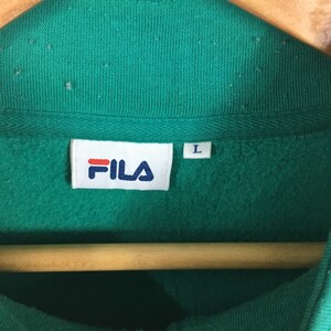 Puede incluir: Una sudadera verde con una etiqueta blanca de Fila y una etiqueta de talla L. La sudadera est&aacute; colgada en una percha de madera.