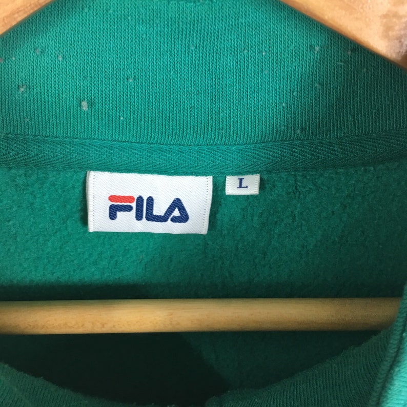 Puede incluir: Una sudadera verde con una etiqueta blanca de Fila y una etiqueta de talla L. La sudadera est&aacute; colgada en una percha de madera.
