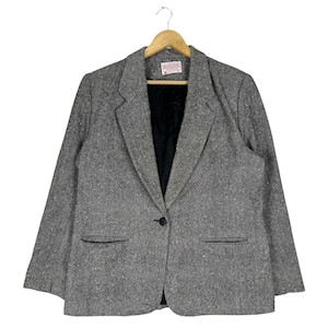 May include: A gray tweed blazer with a single button closure and two flap pockets. The blazer has a black lining and is hanging on a wooden hanger.