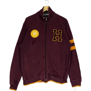May include: A maroon zip-up sweatshirt with a large yellow "H" on the front and a smaller yellow circle with "Tommy Hilfiger" and "Est. 1985" written inside on the left chest. The sweatshirt has yellow stripes on the sleeves.
