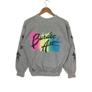 May include: Gray sweatshirt with a colorful graphic on the back that says "Burden Art". The graphic is a rainbow-colored paint splatter with black lettering. The sleeves have a black and white graphic that says "Burden Art" in a stylized font.