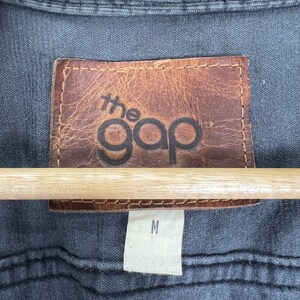 May include: A brown leather patch with the text "the gap" in black lettering. A white tag with the letter "M" is below the patch.