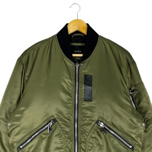 Puede incluir: Una chaqueta bomber verde con detalles de cremallera negra y cierre de cremallera negra. La chaqueta tiene un cuello negro y una etiqueta negra con la palabra "ZARA" en la parte delantera.