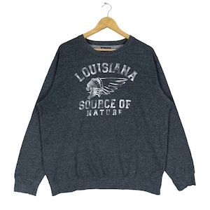 以下が含まれることがあります： 白い羽根と「Louisiana Source of Nature」の文字が書かれたアメリカ先住民の頭飾りの白いグラフィックプリントが施された濃いグレーのスウェットシャツです。