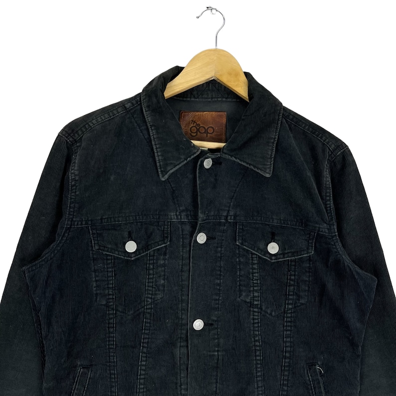 May include: A black corduroy jacket with a button closure and two chest pockets. The jacket has a brown leather label with the text "the gap" on the left side of the chest.