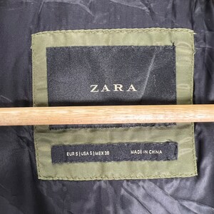 Puede incluir: Una etiqueta negra con texto blanco que dice "ZARA" y "EUR S | USA S | MEX 38" y "MADE IN CHINA". La etiqueta est&aacute; cosida en una tela verde.