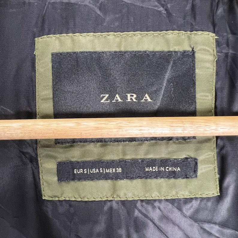 Puede incluir: Una etiqueta negra con texto blanco que dice "ZARA" y "EUR S | USA S | MEX 38" y "MADE IN CHINA". La etiqueta est&aacute; cosida en una tela verde.