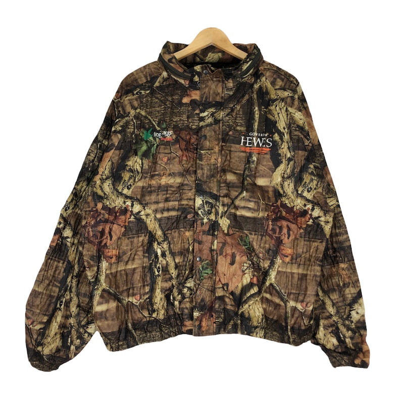 May include: Camouflage jacket with a brown, green, and orange pattern. The jacket has a zipper closure and a button-up flap. The text "GORDON HEWES" is printed on the front of the jacket.