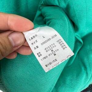 Op de afbeelding: Een groen kledinglabel met witte tekst. Het label toont de maat, het artikelnummer en de stofsamenstelling. Het label zegt "Maat L", "0000399-3038", "Polyester 100%", "Polyester 65% Katoen 35%", "Polyester 65% Katoen 35%" en "Wasinstructies".