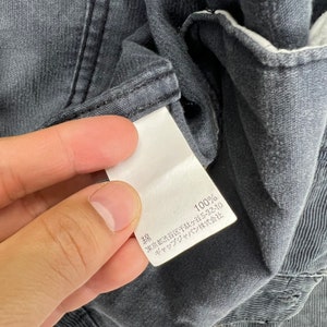May include: A white tag with black text attached to a dark gray garment. The tag reads "綿 100% 東京 都 渋谷 区 千駄ヶ谷 5-32-10  サック イン ジャパン 株式会社"