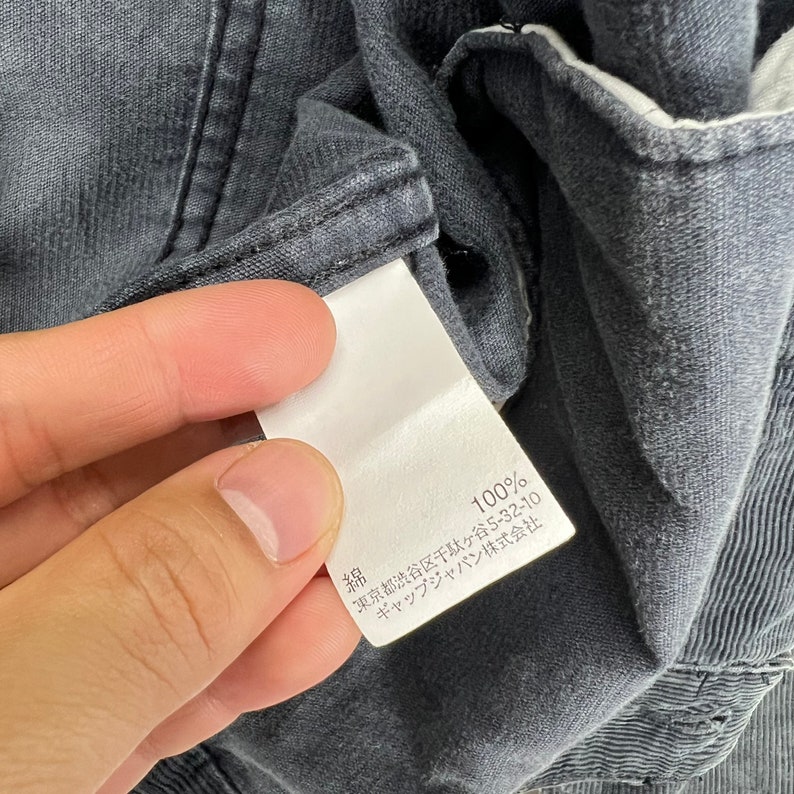 May include: A white tag with black text attached to a dark gray garment. The tag reads "綿 100% 東京 都 渋谷 区 千駄ヶ谷 5-32-10  サック イン ジャパン 株式会社"
