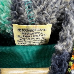Op de afbeelding: Een geel label met zwarte tekst die luidt: "&copy; 2009 BODY GLOVE INTERNATIONAL ALL RIGHTS RESERVED. BODY GLOVE en het Hand Design zijn geregistreerde handelsmerken van Body Glove International, LLC."