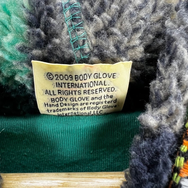 Op de afbeelding: Een geel label met zwarte tekst die luidt: "&copy; 2009 BODY GLOVE INTERNATIONAL ALL RIGHTS RESERVED. BODY GLOVE en het Hand Design zijn geregistreerde handelsmerken van Body Glove International, LLC."