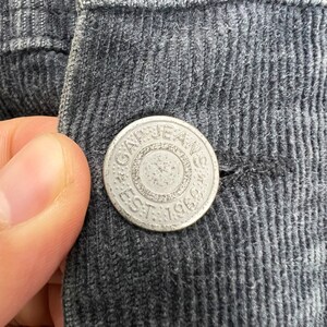 May include: Close-up of a silver button with the text "GAP JEANS EST. 1969" on a dark gray corduroy fabric.