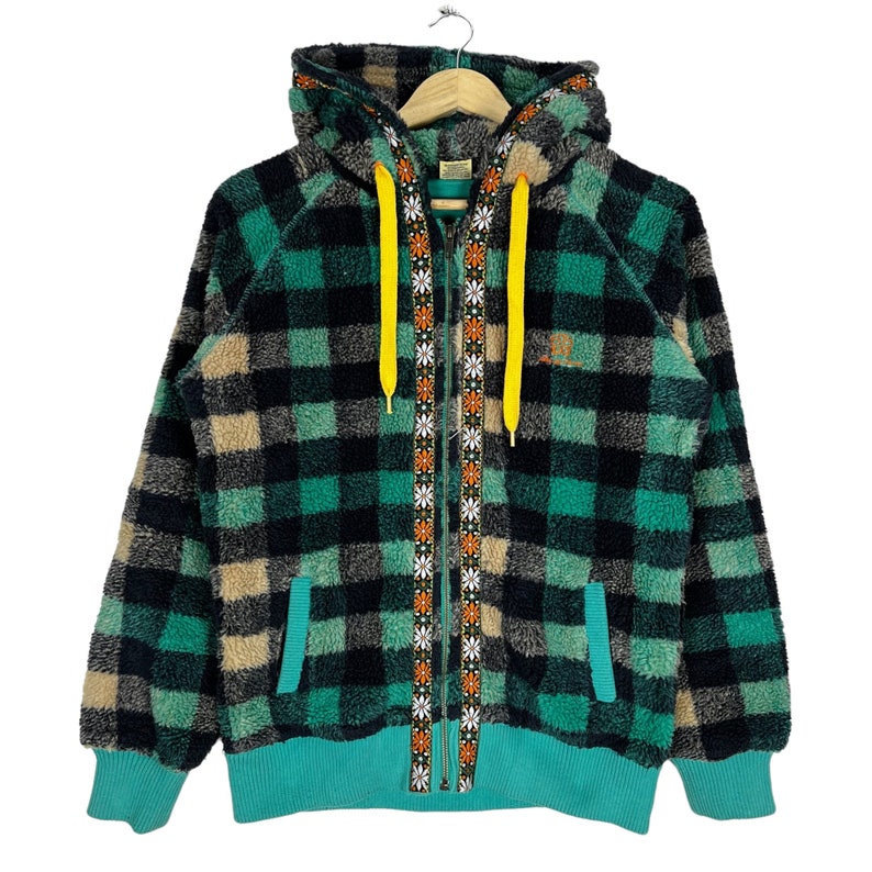Op de afbeelding: Een groene, zwarte en beige geruite fleece hoodie met een gele rits en een rits met een bloemenprint. De hoodie heeft een capuchon en een kangoeroezak.