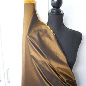 May include: A roll of brown fabric with a golden sheen and a frayed yellow edge. The fabric is draped over a black dress form, with a white background and a glimpse of a white cabinet.