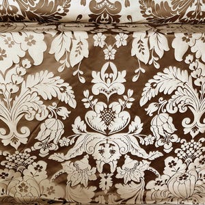 Peut inclure: Gros plan sur un tissu avec un motif floral damassé répété. Le motif présente des fleurs et des feuilles de couleur crème sur un fond marron. Le tissu est plié en haut, révélant le motif.
