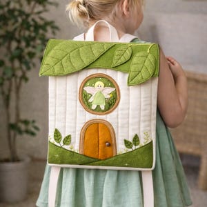 Puede incluir: Una mochila caprichosa con diseño de casa. La tela acolchada blanquecina está adornada con hojas de fieltro verde, una ventana redonda con un hada y una puerta naranja. La mochila tiene correas y un asa blancas.