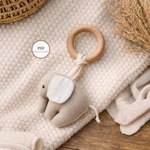 Op de afbeelding: Een beige olifantvormige babyrammelaar met een houten ring en een stoffen oor. De rammelaar is met een crèmekleurige stof aan de ring bevestigd. De afbeelding bevat ook een PDF-naaipatroonlabel.