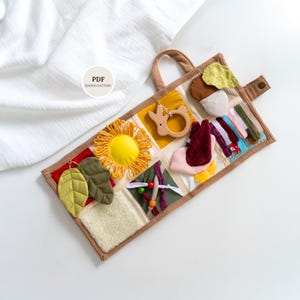 May include: A handmade sensory activity book for babies and toddlers. The book features various textures, colors, and shapes, including a yellow sun, wooden bunny teether, and fabric leaves. The book has a brown corduroy handle and a button closure.