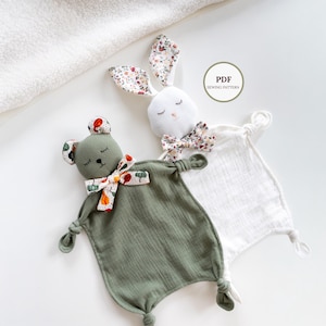 Op de afbeelding: Twee babyknuffels met dierenkoppen. Eén is een witte konijn met bloemenprint oren en strik, de andere is een groene beer met een bloemenprint strik en oren. De dekens zijn vierkant met geknoopte hoeken.