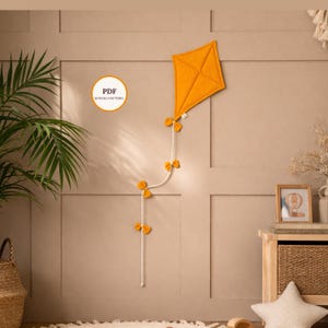 Puede incluir: Una cometa de color amarillo mostaza con un patrón de diamantes está montada en una pared. La cometa está unida a una cuerda blanca con lazos amarillos. Un gráfico circular con el texto "PDF SEWING PATTERN" también está en la pared.
