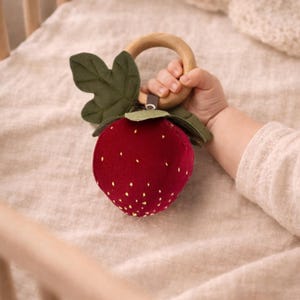 Könnte beinhalten: Ein Babyrassel in Erdbeerform. Die rote Plüschfrucht hat gelbe Samendetails und grüne Blätter. Sie hat einen Holzring zum Halten. Das Spielzeug liegt auf einer beigen Oberfläche.