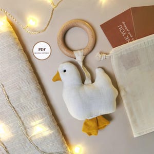 May include: A handmade white fabric duck toy with a wooden ring and yellow beak and feet. The duck is attached to the ring with a white fabric strap. A small white drawstring bag and a card that says "THANK YOU" are also in the image. The text "PDF SEWING PATTERN" is in a circle.