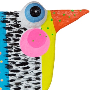 Hautnah-Aufnahme des Kopfes einer handgefertigten Vogel Figur aus Pappmaché. Im Fokus stehen das markante, hervorstehende blaue Auge, der gepunktete orange Schnabel und die rosa Wange. Das Foto zeigt die feine Textur der moderne naive Kunst Skulptur sowie die aufwendige Handbemalung mit gelben, blauen und schwarz-weißen Mustern. Dieser Fantasievogel als Unikat ist ein faszinierender Blickfang für Wohn- und Arbeitsräume.