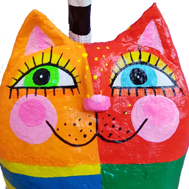 Eine farbenfrohe, skurrile Katzenskulptur mit geteiltem Gesichtsdesign. Eine Seite ist orange, die andere rot, mit großen, ausdrucksstarken Augen in Grün und Blau. Rosa Wangen und eine kleine rosa Nase vervollständigen das Gesicht. Die Katze hat einen schwarz-weiß gestreiften Schwanz.