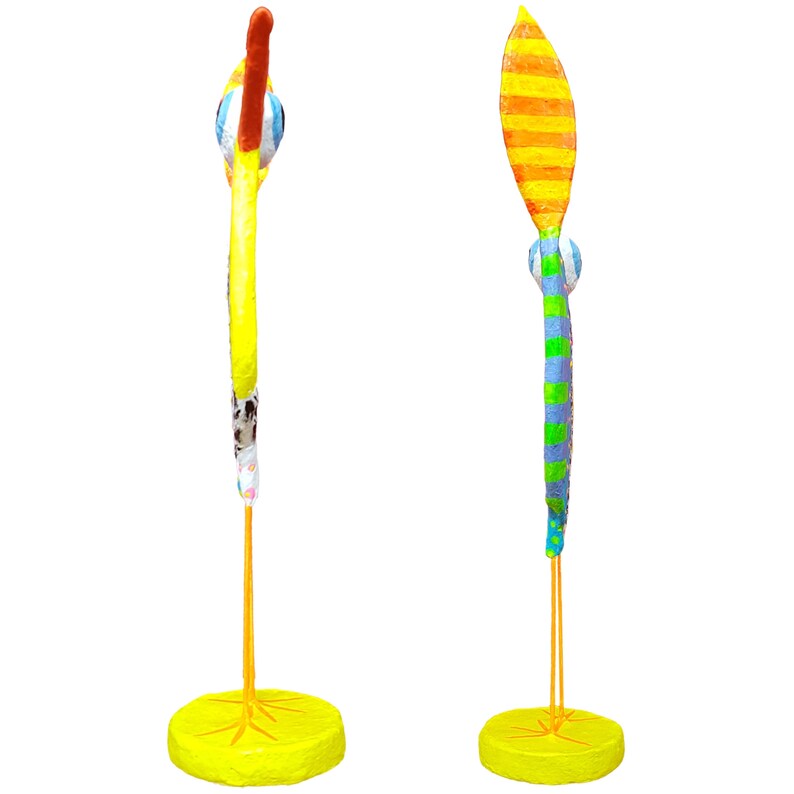 Zwei Seitenansichten des Profils einer handgefertigten Vogel Figur aus Pappmaché. Links die gelbe Vorderseite mit markanter rosa Wange und dem orangefarbenen Schnabel. Rechts die blaue Rückseite mit bunten Punkten, dem orange-gelb gestreiften Schwanz und dem grünen Streifenmuster. Die Skulptur im Stil der modernen naiven Kunst steht auf grazilen orangefarbenen Beinen und einem gelben Sockel. Ein faszinierender Fantasievogel als Unikat für Wohn- und Arbeitsräume.