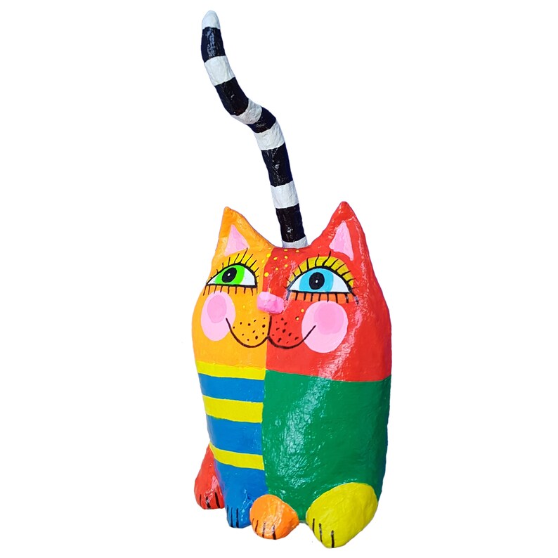Eine skurrile, farbenfrohe Katzenskulptur. Die Katze ist in leuchtenden Farben von Orange, Gelb, Rot, Grün und Blau bemalt. Der Schwanz ist schwarz-weiß gestreift. Die Katze hat ein lächelndes Gesicht mit großen Augen und rosigen Wangen.